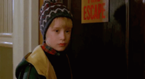 A Young Boy Wearing A Beanie Is Standing In Front Of A Door With A Sign That Says Escape . GIF