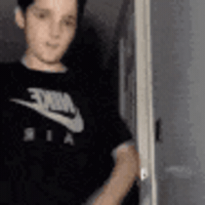 A Young Boy Wearing A Black Nike T-Shirt Is Standing In Front Of A Door . GIF