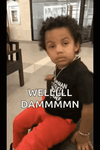 A Young Boy Wearing A Black Shirt And Red Pants Is Sitting On A Chair And Making A Funny Face . GIF