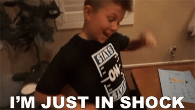 A Young Boy Wearing A Black Shirt That Says I 'M Just In Shock Is Standing In Front Of A Desk . GIF