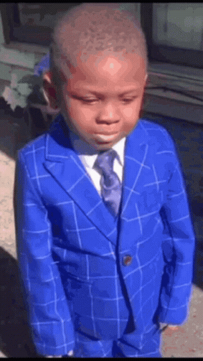 A Young Boy Wearing A Blue Suit And Tie Is Standing In Front Of A Window . GIF
