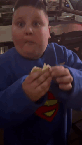 A Young Boy Wearing A Blue Superman Sweatshirt Is Eating Something GIF