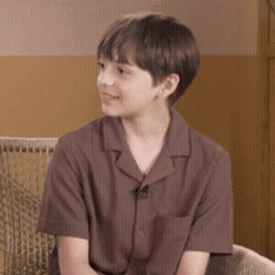 A Young Boy Wearing A Brown Shirt Is Sitting On A Chair . GIF