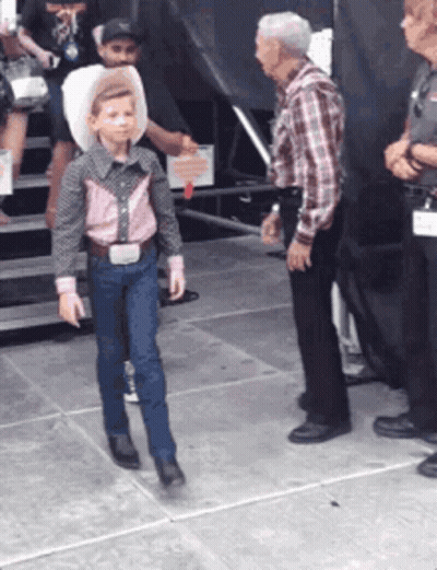A Young Boy Wearing A Cowboy Hat And Suspenders Is Walking On The Sidewalk GIF