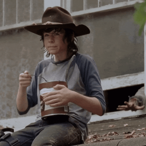 A Young Boy Wearing A Cowboy Hat Is Eating From A Bucket Of Food GIF