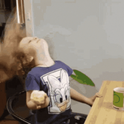 A Young Boy Wearing A Donald Duck Shirt Is Sitting At A Table GIF