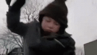 A Young Boy Wearing A Hooded Jacket And A Hat Is Throwing Snow In The Air . GIF