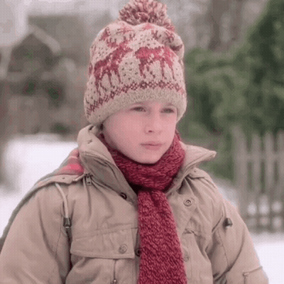 A Young Boy Wearing A Knitted Hat And Scarf Stands In The Snow GIF