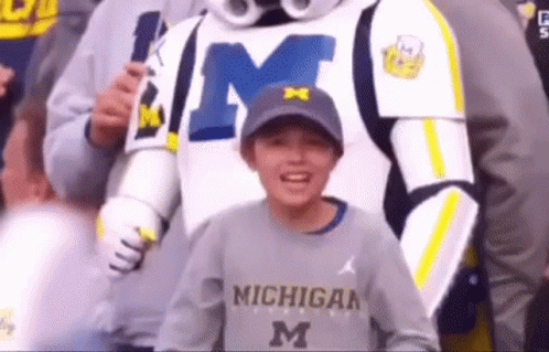 A Young Boy Wearing A Michigan Sweatshirt Is Standing Next To A Mascot GIF