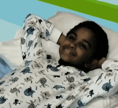 A Young Boy Wearing A Pajama Top With Dinosaurs On It Is Laying On A Bed GIF