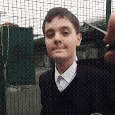 A Young Boy Wearing A School Uniform Stands In Front Of A Fence GIF