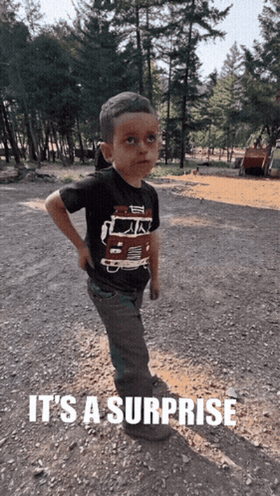 A Young Boy Wearing A Shirt That Says ' It 'S A Surprise ' Is Standing In The Dirt GIF