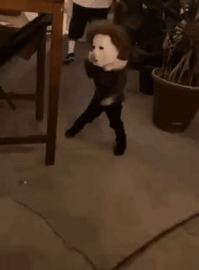 A Young Boy Wearing A White Mask Is Standing Next To A Potted Plant . GIF