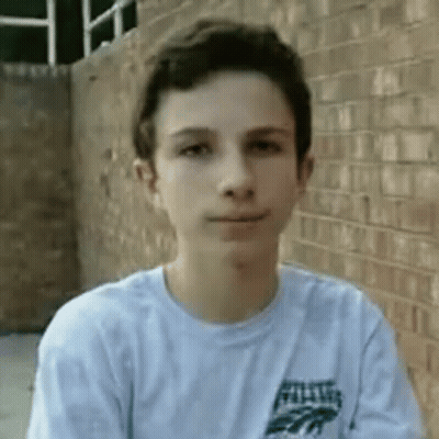 A Young Boy Wearing A White Shirt Is Standing In Front Of A Brick Wall And Looking At The Camera . GIF