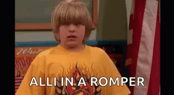 A Young Boy Wearing A Yellow Shirt Is Standing In Front Of An American Flag And Says `` All In A Romper '' . GIF