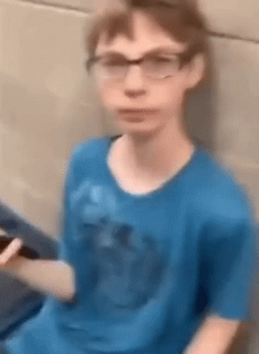 A Young Boy Wearing Glasses And A Blue Shirt Is Sitting On A Couch Holding A Cell Phone . GIF