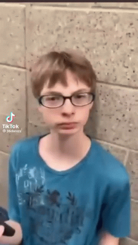 A Young Boy Wearing Glasses Is Standing In Front Of A Brick Wall . GIF