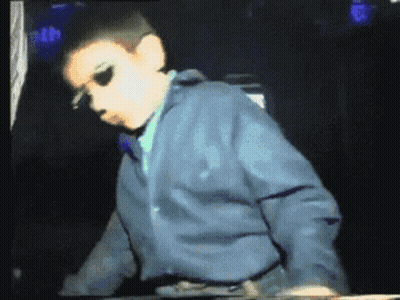 A Young Boy Wearing Sunglasses And A Blue Jacket Stands In A Dark Room GIF