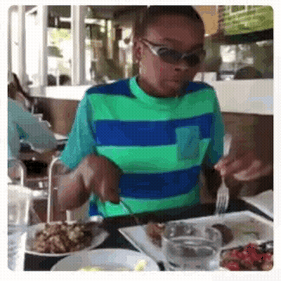 A Young Boy Wearing Sunglasses Is Sitting At A Table With Plates Of Food . GIF