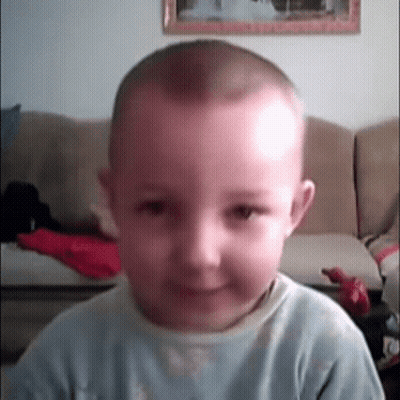 A Young Boy With A Shaved Head Is Smiling In Front Of A Couch GIF