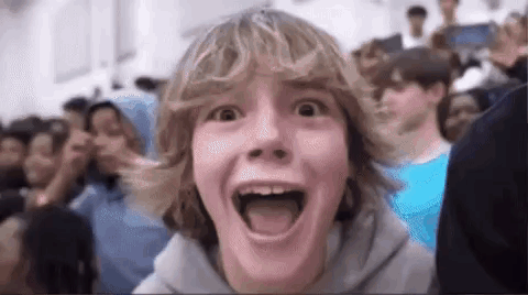 A Young Boy With Blonde Hair Is Making A Funny Face In Front Of A Crowd Of People . GIF