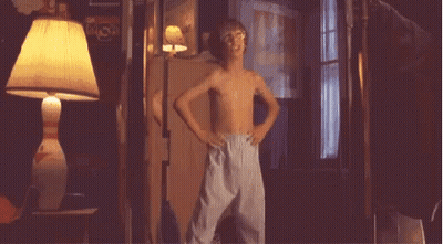 A Young Boy Without A Shirt Is Standing In A Dark Room With A Lamp . GIF