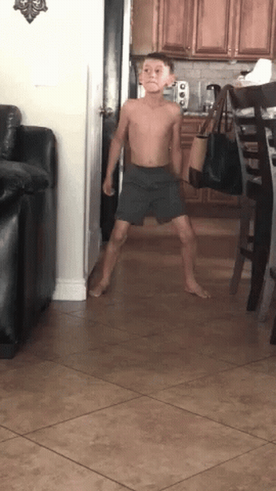 A Young Boy Without A Shirt Is Standing In A Living Room . GIF