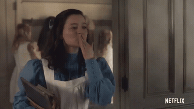 A Young Girl In A Blue Dress And White Apron Is Standing In Front Of A Door Holding A Book And Waving . GIF