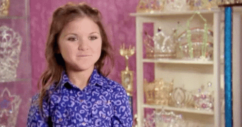A Young Girl In A Blue Shirt Is Standing In Front Of A Shelf Filled With Crowns And Tiaras . GIF