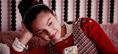 A Young Girl In A Red Sweater Is Laying On A Bed Looking At Her Phone . GIF