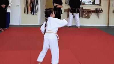 A Young Girl In A White Karate Uniform Is Standing On A Red Mat In A Gym . GIF