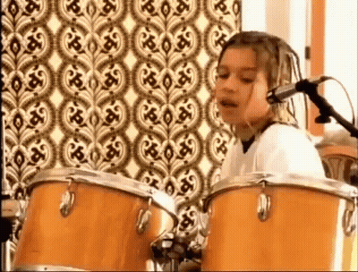 A Young Girl Is Playing Drums And Singing Into A Microphone In Front Of A Patterned Wall . GIF