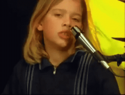 A Young Girl Is Singing Into A Microphone With A Yellow Background Behind Her . GIF