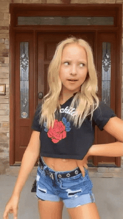 A Young Girl Wearing A Black Crop Top And Shorts Is Standing In Front Of A Door . GIF