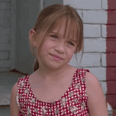 A Young Girl Wearing A Red And White Plaid Dress Stands In Front Of A Brick Wall GIF