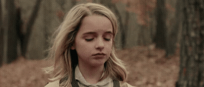 A Young Girl With Blonde Hair Is Standing In The Woods With Her Eyes Closed And Her Hair Blowing In The Wind . GIF