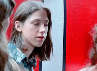 A Young Girl With Long Hair Is Standing In Front Of A Red Wall And Looking At The Camera . GIF