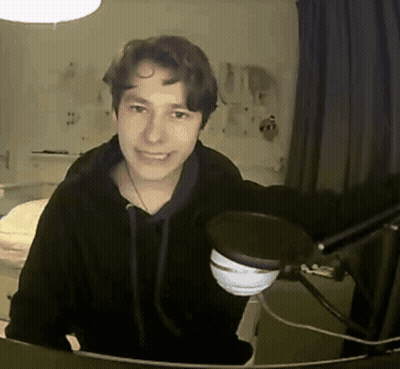 A Young Man In A Black Hoodie Stands In Front Of A Microphone And Smiles GIF