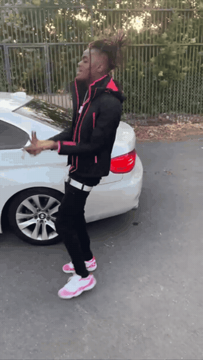 A Young Man In A Black Jacket And Pink Shoes Is Standing In Front Of A White Car GIF