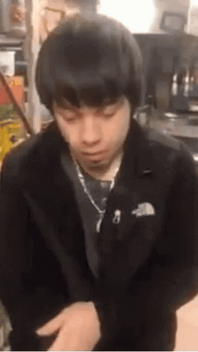 A Young Man In A Black Jacket And Tie Is Sitting In A Kitchen . GIF