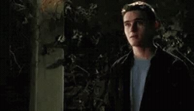 A Young Man In A Black Jacket And White Shirt Is Standing In Front Of A Tree At Night . GIF