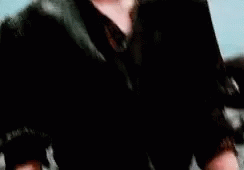 A Young Man In A Black Jacket Is Standing In Front Of A Blue Background . GIF