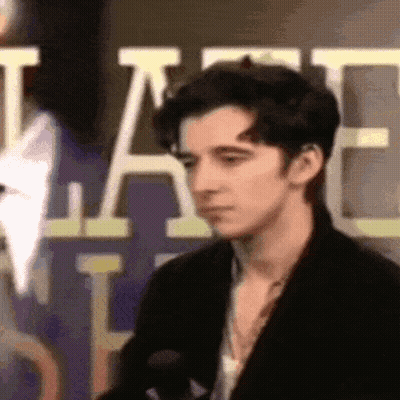 A Young Man In A Black Jacket Is Standing In Front Of A Sign That Says ' A ' On It . GIF