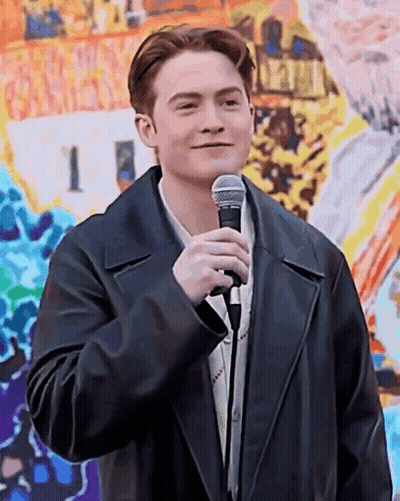 A Young Man In A Black Leather Jacket Is Holding A Microphone In Front Of A Painting . GIF