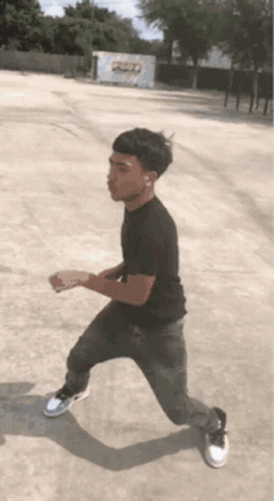 A Young Man In A Black Shirt And Grey Jeans Is Standing In A Parking Lot GIF