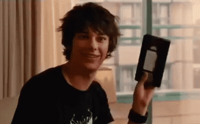 A Young Man In A Black Shirt Is Holding A Vhs Tape In His Hand . GIF
