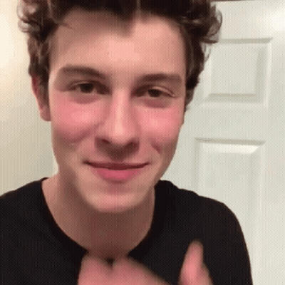 A Young Man In A Black Shirt Is Smiling And Giving A Thumbs Up While Standing In Front Of A Door . GIF