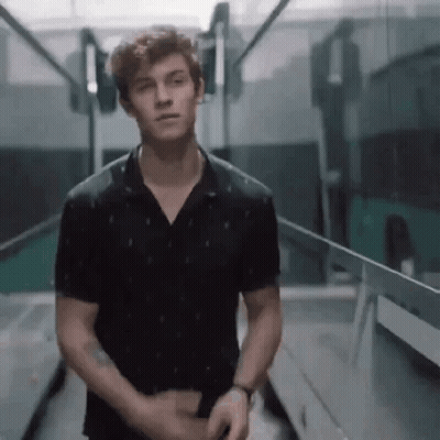 A Young Man In A Black Shirt Is Standing In A Hallway Looking At The Camera . GIF