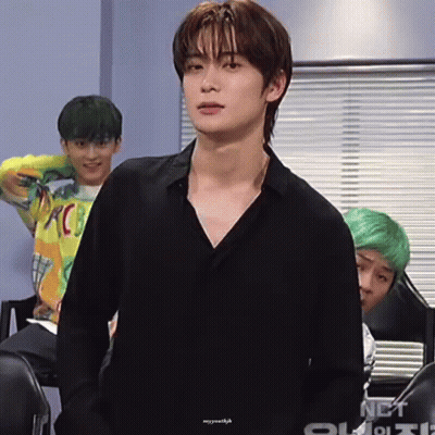A Young Man In A Black Shirt Is Standing In Front Of A Window With A Group Of Young Men Behind Him . GIF