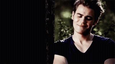 A Young Man In A Black Shirt Is Standing Next To A Tree And Smiling . GIF